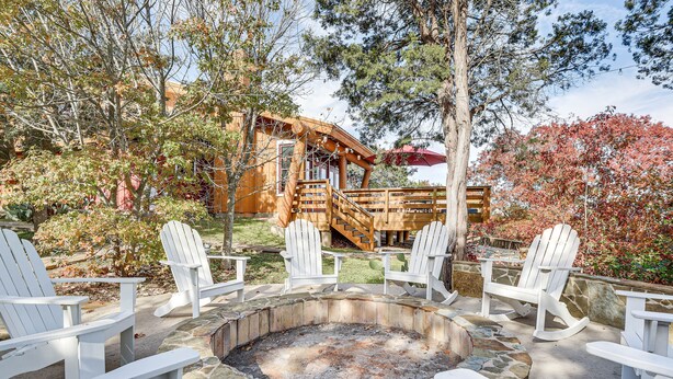 A stone fire pit surrounded by white Adirondack chairs, a wooden deck, and a rustic building with a wooden staircase. - Lake Whitney Cabin w/ Hot Tub & Fire Pit! (Morgan)