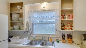 A kitchen with a double sink, a window with a lace curtain, and shelves with dishes and kitchen items. - Convenient, Pet-Friendly Rochester Vacation Rental (Rochester)