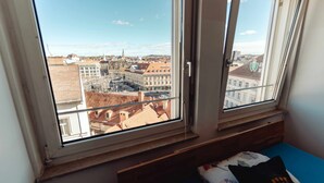 Ett rum med ett stort fönster med utsikt över stadsbilden, en säng med en blå madrass och en kudde med text på.