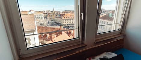 Une chambre dotée d’une grande fenêtre donnant sur un paysage urbain, d’un lit avec un matelas bleu et d’un oreiller orné d’un motif textuel.