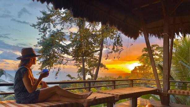 A person relaxing on a wooden bench with a view of the sunset over the ocean, a wooden table with a basket of fruit, and a thatched roof structure. - Finca Exotica ecolodge (Puerto Jiménez)