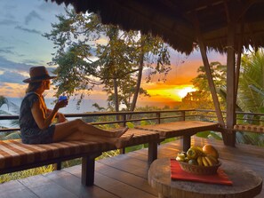 A person relaxing on a wooden bench with a view of the sunset over the ocean, a wooden table with a basket of fruit, and a thatched roof structure. - Finca Exotica ecolodge (Puerto Jiménez)