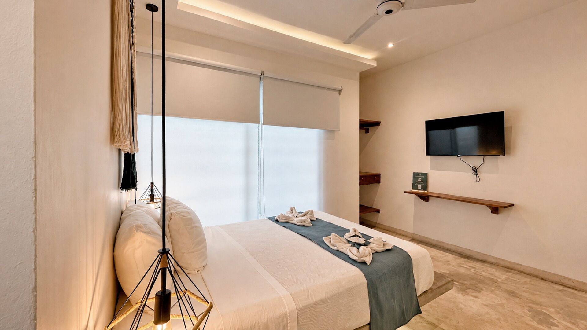 A modern bedroom with a bed, a hanging light fixture, a TV mounted on the wall, and a window with blinds.