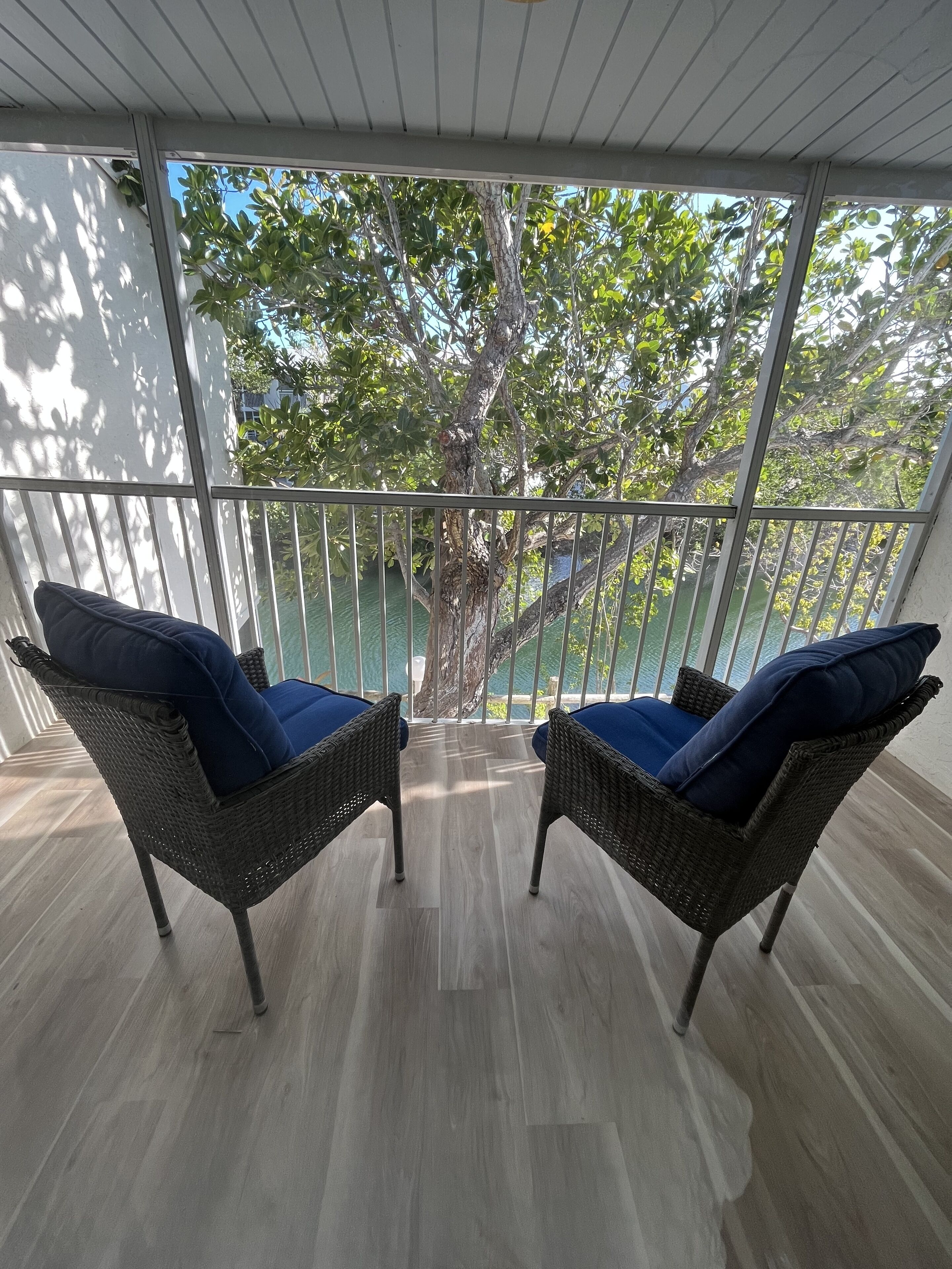A balcony with wicker chairs and cushions, a view of a tree, and a railing.