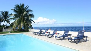 Zona de piscina con sillones de descanso y palmeras, con vistas al océano.