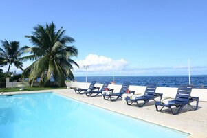 A poolside area with lounge chairs and palm trees, overlooking the ocean.