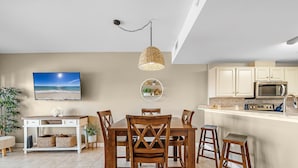 Un área de comedor con una mesa y sillas de madera, un televisor en la pared y una cocina con armarios y electrodomésticos.