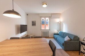 A modern living room with a wooden dining table, a blue sofa, a TV, and a window with curtains. - HouSmart Corte Galluzzi 7/2 (Bologna)