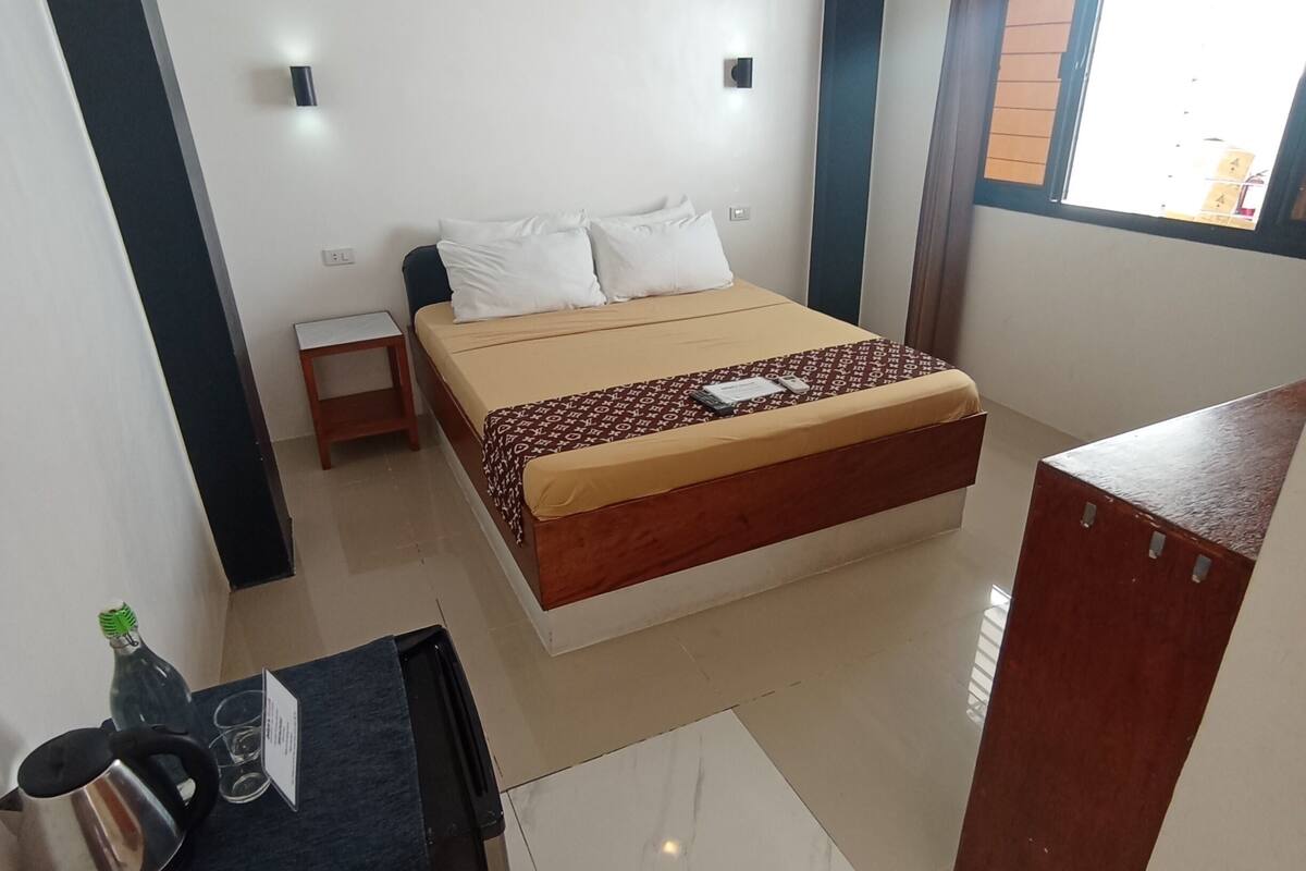 A bedroom with a bed, a small table with a kettle, a cabinet, a ceiling fan, and an air conditioning unit.