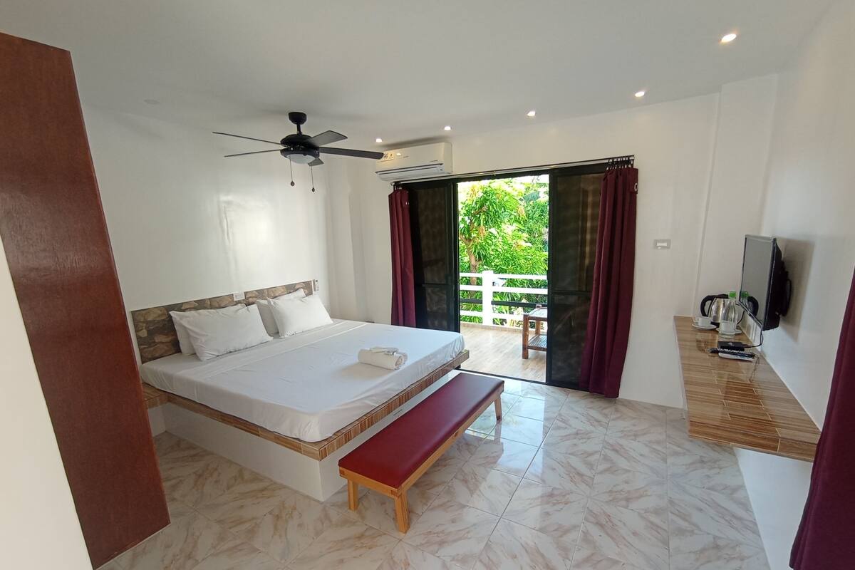 A bedroom with a bed, a bench, a ceiling fan, a TV, and a view of greenery outside.