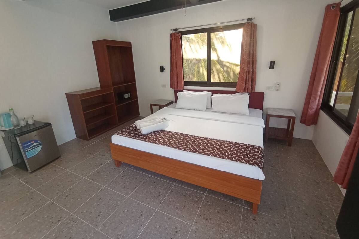 A bedroom with a bed, a wooden wardrobe, a small table, a ceiling fan, and a window with curtains.