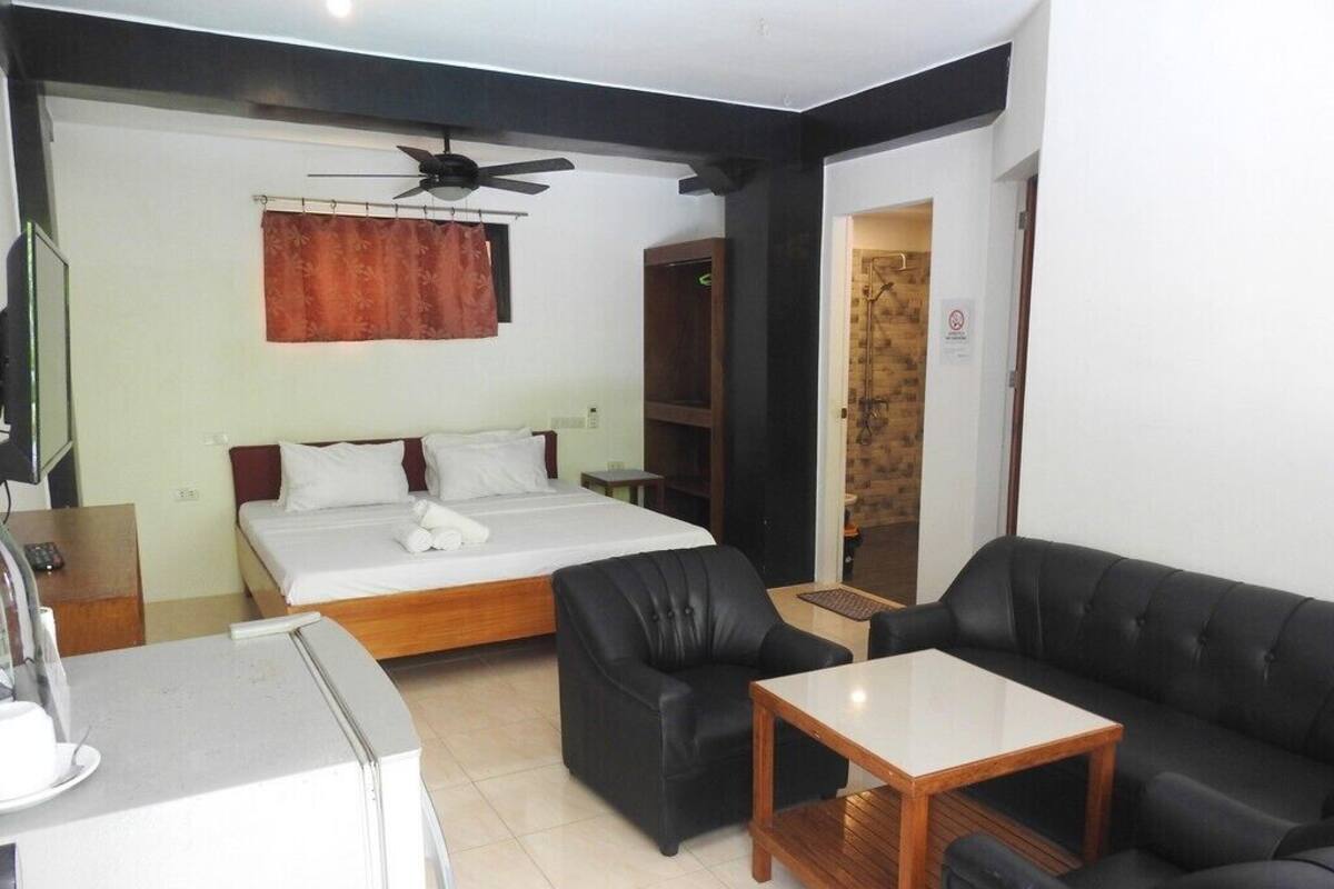 A hotel room with a bed, two black leather sofas, a small wooden table, a ceiling fan, and a shower area.