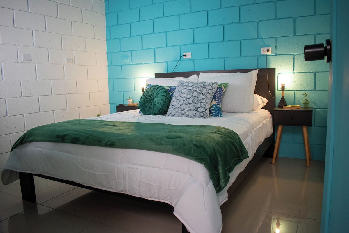 Una cama con cubrecama verde y blanco, una mesita de noche de madera con lámpara y una pared de ladrillo azul.
