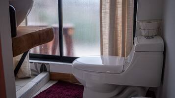 A small bathroom with a toilet, a wooden shelf, a window with curtains, and a red rug.
