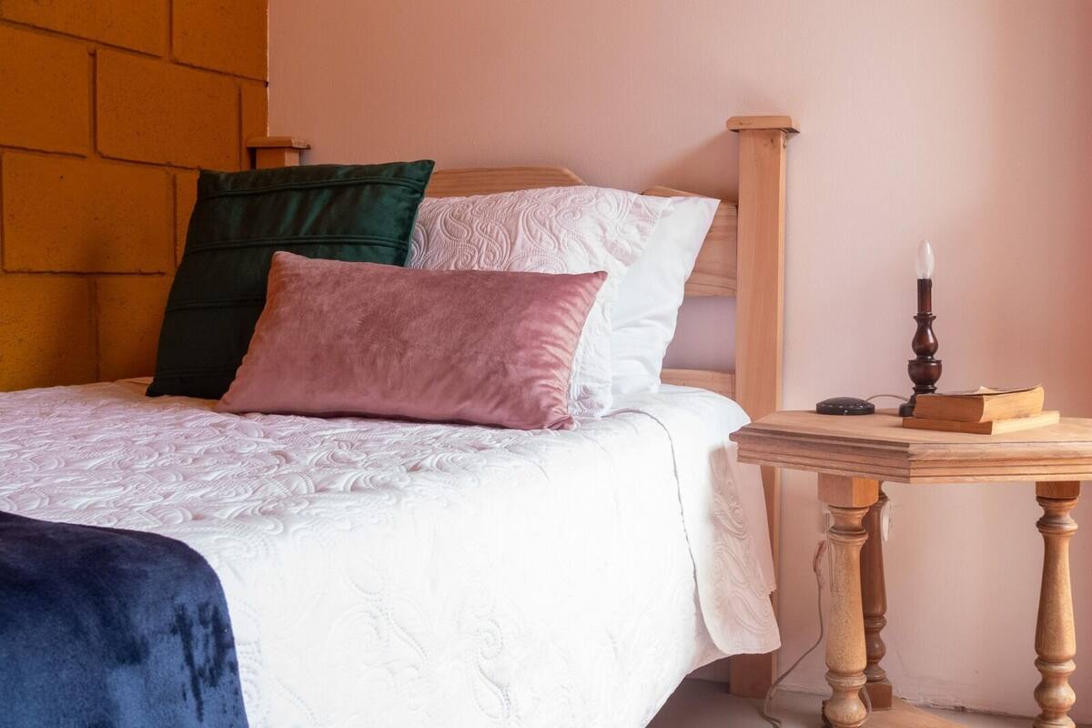 Una cama bien hecha con edredón blanco y varios almohadones, una mesita de noche de madera con un libro y una vela, y una pared de ladrillo al fondo.