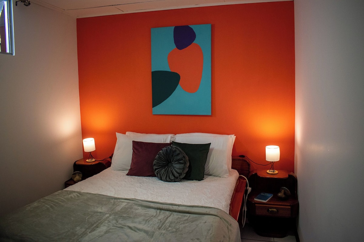 Una cama bien hecha con sábanas blancas y un plaid verde, flanqueada por dos mesitas de noche con lámparas, en una habitación con una pared de acento naranja y un cuadro abstracto moderno.