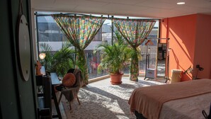 A bedroom with a large bed, a desk with a chair, a potted plant, and a view through a window with curtains. - Lolas Hostal (San José)