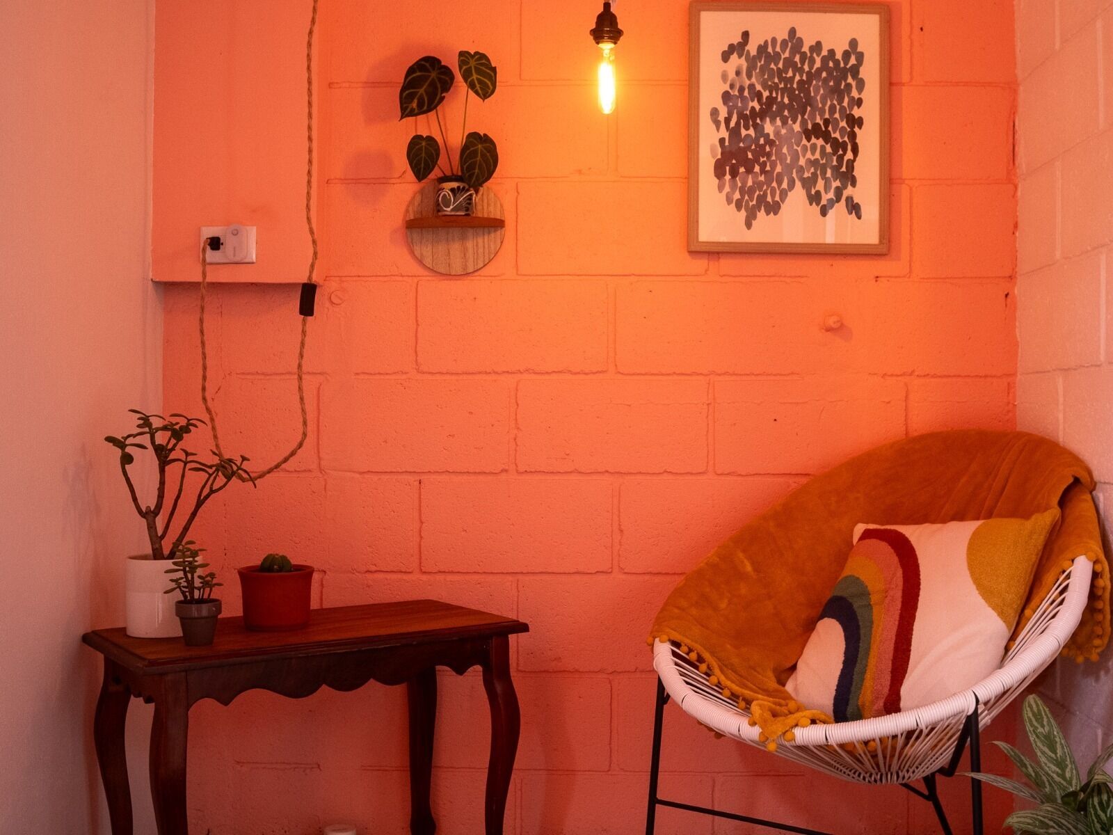 A room with a brick wall, a wooden table with potted plants, a wicker chair with a colorful cushion, a wall-mounted light fixture, and a framed artwork.