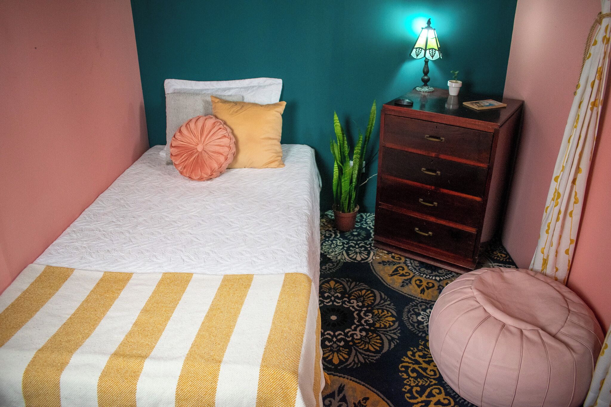 Una cama con un edredón a rayas, una mesita de noche con una lámpara, una planta en maceta y una alfombra estampada.