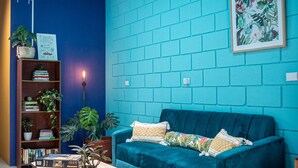 A room with a blue brick wall, a teal sofa, a wooden coffee table, and a bookshelf with books and plants. - Lolas Hostal (San José)