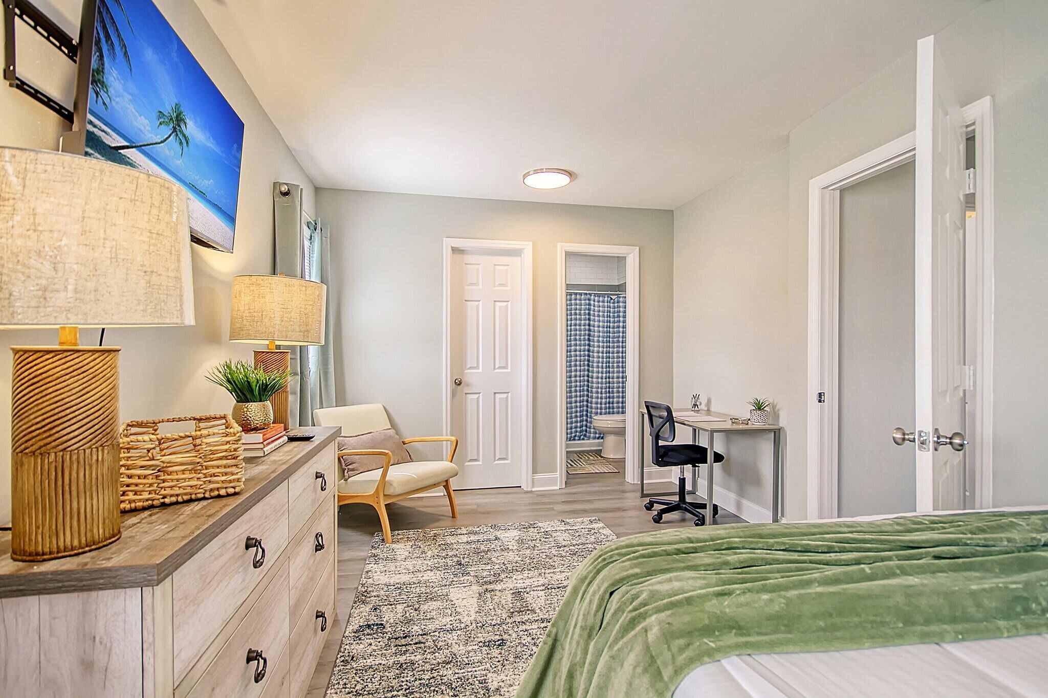 A bedroom with a bed, dresser, desk, chair, and a view of the bathroom.