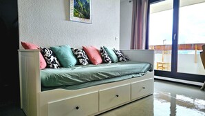 A modern room with a white sofa bed, a small table, and a window with curtains. - Appartement Aldeïa - Welkeys (Huez)