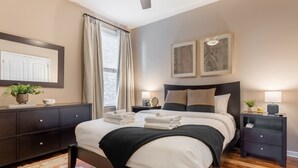 A modern bedroom with a large bed, dark wood headboard, bedside tables with lamps, a mirror, and a view of the outside through a window with curtains. - Elegant Home with Large Yard (Nashville)