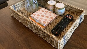 A woven tray on a table with a vase of green foliage, a notebook, two decorative tins, and a remote control. - Stylish Family Friendly Bungalow, Tvs, Royal Beds (Hinesville)