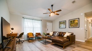 A living room with a brown leather sofa, a coffee table, and a desk with a lamp. There is a ceiling fan, a window with sheer curtains, and two framed pictures on the wall. - Stylish Family Friendly Bungalow, Tvs, Royal Beds (Hinesville)