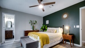 A bedroom with a bed, bedside tables, a bench, a ceiling fan, a mirror, a plant, and a wall-mounted light fixture. - Stylish Family Friendly Bungalow, Tvs, Royal Beds (Hinesville)