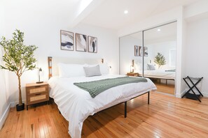 A modern bedroom with a large bed, wooden flooring, a nightstand with a lamp, and a potted plant. - 3-Bed Apartment Near Disney with WiFi, AC • Kid Friendly (Santa Ana)