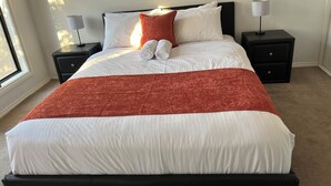 A neatly made bed with white linens and a red throw blanket, flanked by two nightstands with lamps, in a room with a large window. - Tasha's Apartments on Kerry (Athol Park)