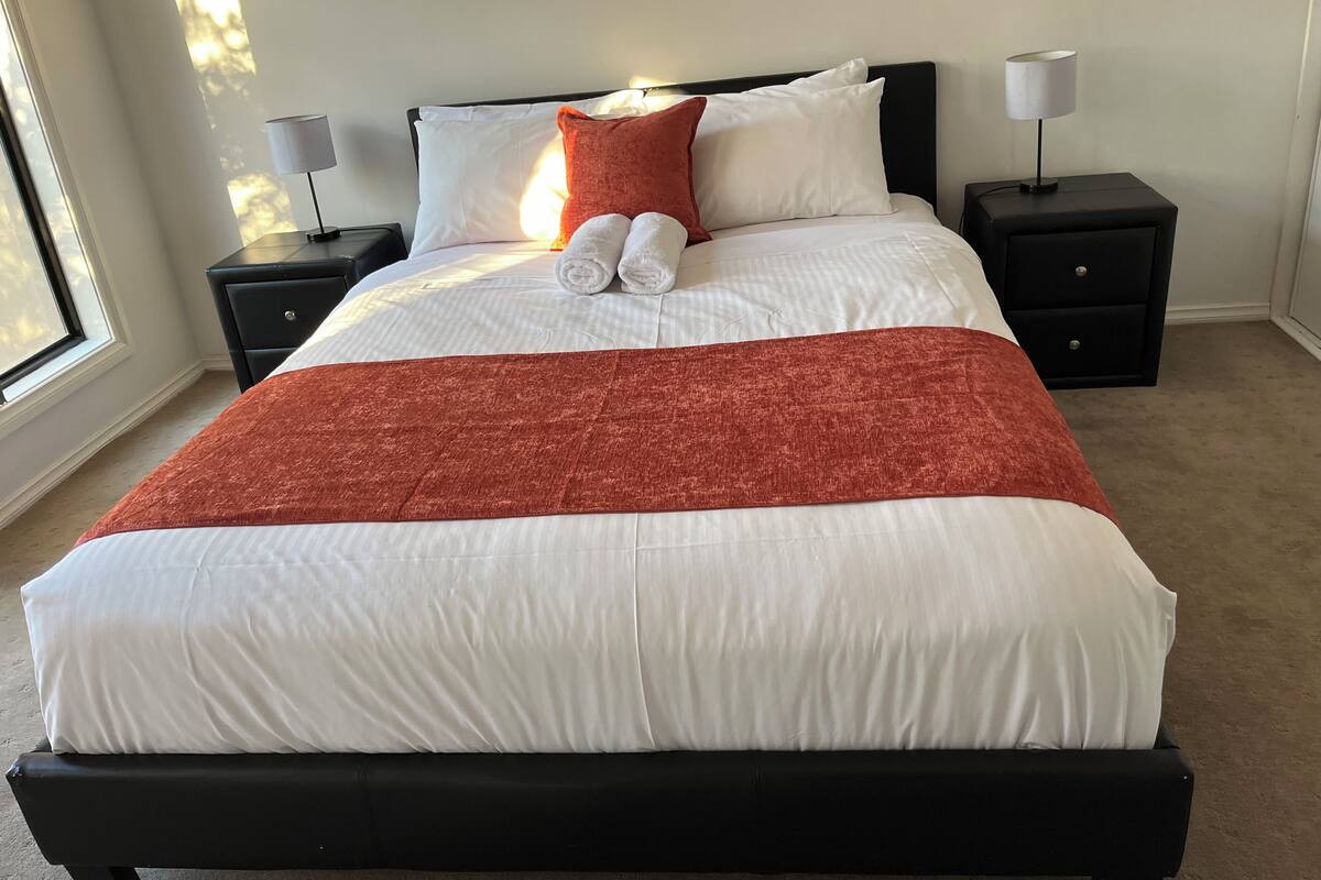 A neatly made bed with white linens and a red throw blanket, flanked by two nightstands with lamps, in a room with a large window.