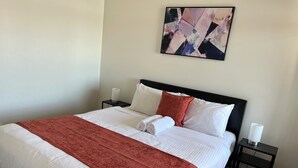 A neatly made bed with white and red bedding, a black headboard, and a small bedside table with a lamp. - Tasha's Apartments on Kerry (Athol Park)