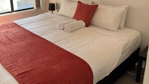 A neatly made bed with white and red bedding, a wall-mounted artwork, and a view of a parked car through a window. - Tasha's Apartments on Kerry (Athol Park)