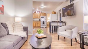 A modern living room with a grey sofa, a round coffee table with a plant, and a grey armchair. A TV is mounted on the wall, and a kitchen is visible in the background. - Clean and comfortable with astounding views of Pikes Peak.  (Colorado Springs)
