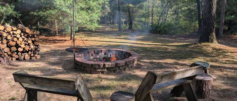 A campfire surrounded by a stone fire pit in a forested area with a stack of firewood and two wooden benches.