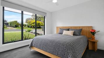 A modern bedroom with a large window, a bed with striped bedding, a wooden headboard, and a small side table with a vase.