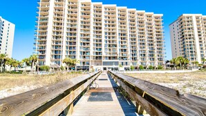 A wooden boardwalk leading to a large, multi-story hotel with balconies and a clear blue sky. - Beachfront 1BR + Bunk Alcove @ Phoenix East • Sleeps 6 • Amazing Views & Resort Amenities (Orange Beach)