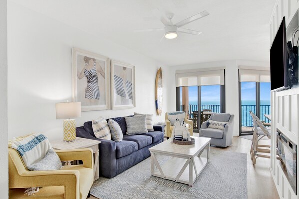 Un salon moderne avec un canapé, des fauteuils, une table basse et un téléviseur. Des œuvres d’art encadrées ornent le mur, et de grandes fenêtres offrent une vue sur la mer.