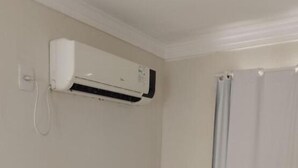 A small, white room with a white air conditioning unit mounted on the wall, a small white refrigerator, and a shower area with a white curtain. - Hotel California (Goiânia)