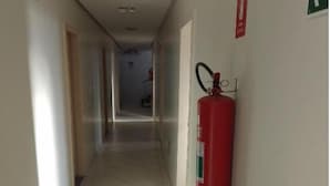 A corridor with a fire extinguisher mounted on the wall, a fire exit sign, and a tiled floor. - Hotel California (Goiânia)
