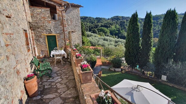 A stone building with a green door, outdoor seating with chairs and a table, potted plants, and a view of a lush green landscape with trees. - Beautiful  villa with WIFI, private pool, TV, terrace and panoramic view, close to Lucca (Chiatri)