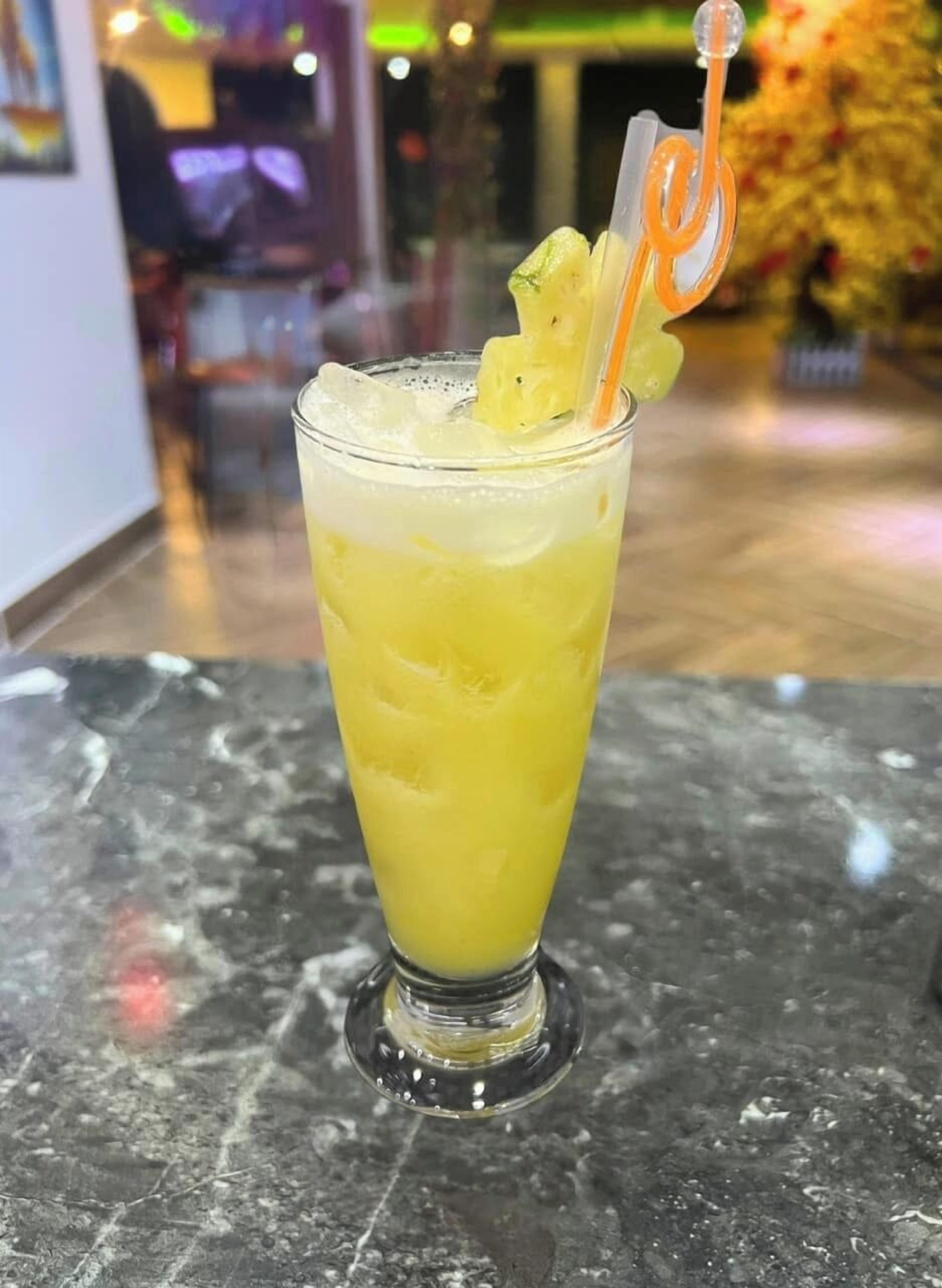 A tall glass of yellow beverage with ice cubes and a straw, placed on a marble surface.