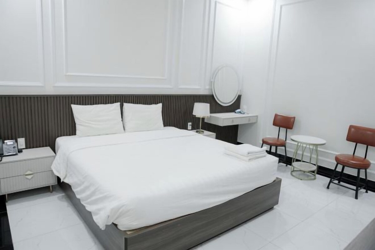Habitación de hotel moderna con una cama grande, mesitas de noche, un tocador con espejo y dos sillas.