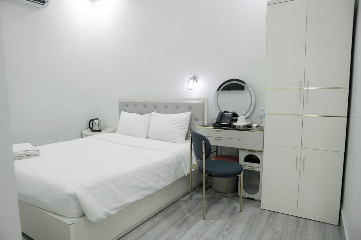 Une chambre d’hôtel comprenant un lit, un bureau avec une chaise, une armoire, un miroir et une petite poubelle.
