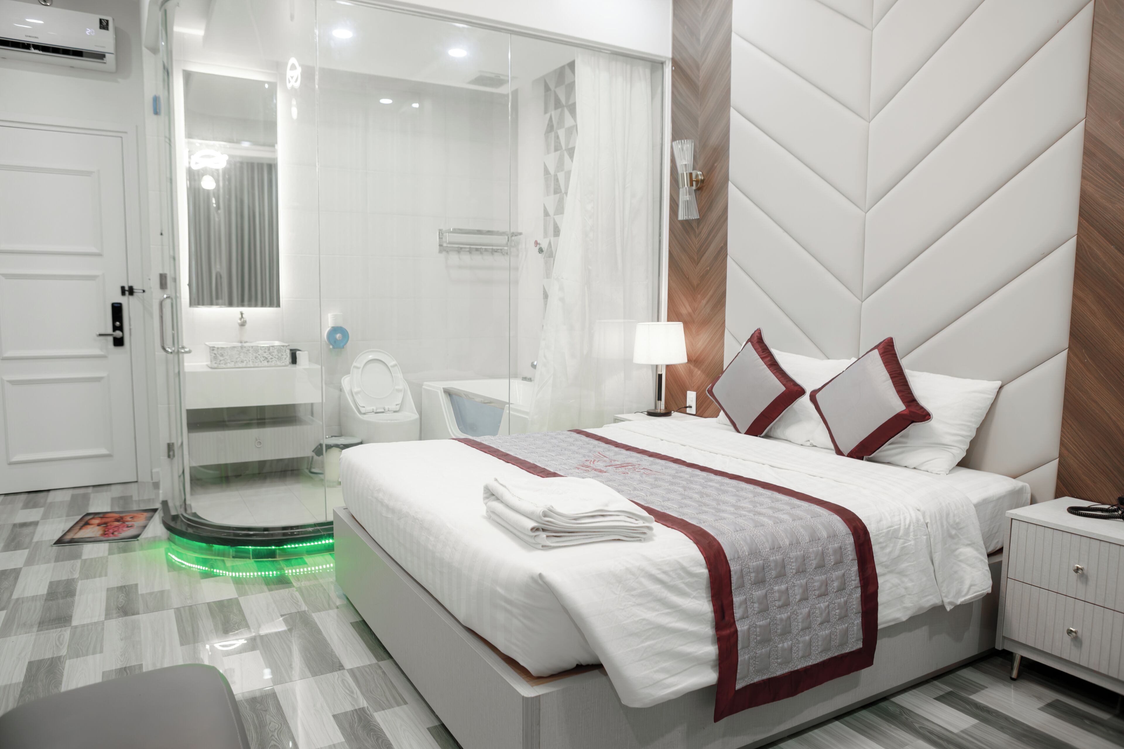 A modern hotel room with a large bed, a glass shower enclosure, a vanity with a mirror, and a bedside table with a lamp.