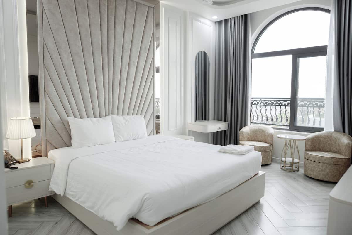 Une chambre d’hôtel moderne avec un grand lit, une table de chevet avec une lampe, une petite table ronde, deux fauteuils et une vue sur la ville par une grande fenêtre.