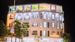 Un hotel de varios pisos con balcones, un cartel que dice 'TAN HANH HOTEL' y un evento de 'GRAN INAUGURACIÓN'.