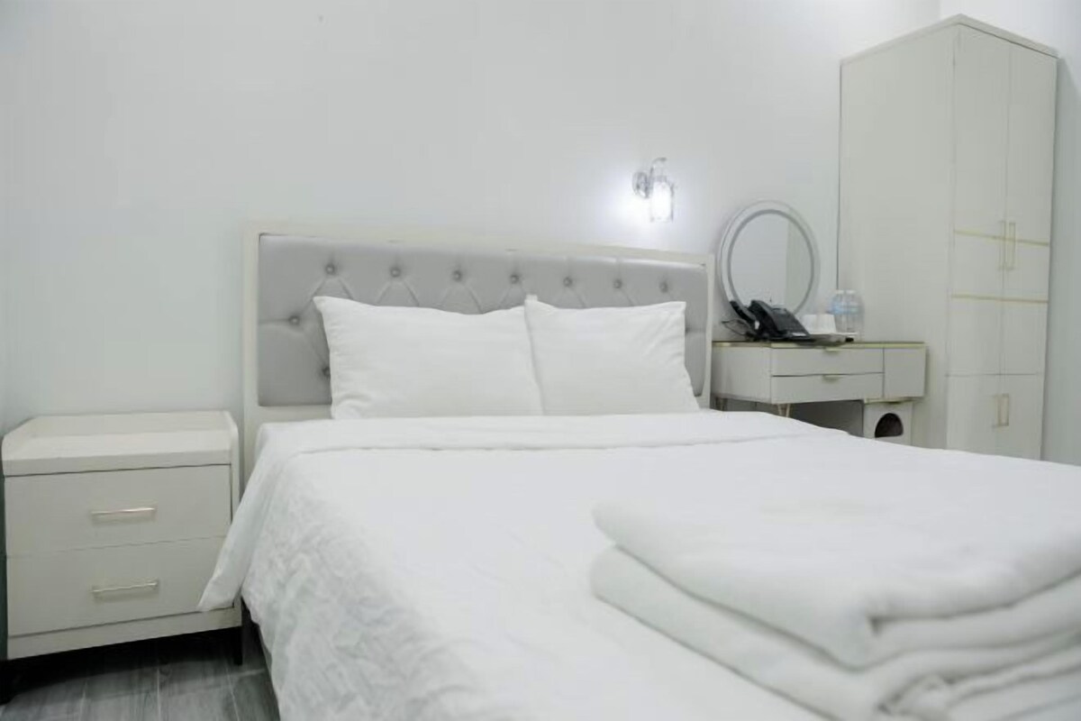 Una cama bien hecha con sábanas blancas, una mesita de noche con lámpara y un armario en una habitación de hotel.